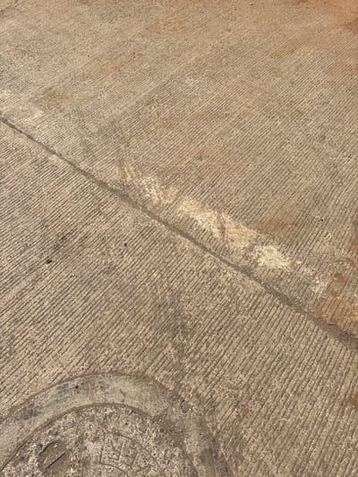 This image displays a section of a grooved concrete surface in Ambernath, India. The light brown concrete features distinct parallel diagonal grooves. A straight, dark linear joint traverses the frame from the upper right to the lower left. Irregular patches of lighter-colored residue or staining are present along and near this joint. A partial view of a circular, textured metal utility access cover is visible in the lower left corner. The surface exhibits minor debris and appears worn. No human subjects, animals, or active events are depicted.