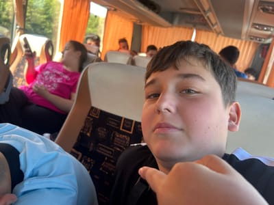 The image depicts the interior of a bus, possibly in Polsko Padarevo, Bulgaria, showing passengers. In the foreground, a young boy is taking a selfie, looking directly at the camera with a neutral expression. He is wearing a black t-shirt with white stripes on the sleeves. His right hand is raised, with his index finger extended towards the lens. Behind him, other passengers are visible. To the left, a girl in a bright pink t-shirt is looking at her phone, and further back, other individuals are blurred in the background, some with their faces partially obscured. The bus features orange curtains on the windows and light-colored seats. The overall lighting suggests it is daytime, with sunlight filtering through the windows.