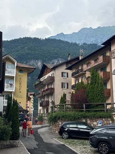 This image depicts a street scene in a mountainous region, likely Molveno, Italy, given the text on a building. The orientation of the image is vertical, requiring rotation to view correctly.

In the foreground, two people are walking along a paved street. The person on the left is wearing an orange top and what appear to be khaki shorts. They are carrying a red bag in their left hand. The person on the right is wearing a red top and dark pants and is holding a red umbrella. They are walking away from the camera. Behind them, a dark-colored car is parked on the right side of the street.

The scene is dominated by buildings and natural scenery. A multi-story building with balconies and greenery climbing its facade is prominent. Further back, a rugged, tree-covered mountainside rises under a cloudy sky. The architecture suggests a European setting, with the building's balconies and window shutters.

On the yellow facade of one building, the text "HOTEL EUROPA MOLVENO" is clearly visible, along with smaller text that appears to read "Lake & Mountain". This confirms the location as Molveno and indicates the presence of a hotel. Another building further down the street has visible signage for "Illy".

The weather appears to be overcast, with a soft, diffused light, suggesting it might be a cloudy day or early morning/late afternoon. The overall atmosphere is serene, with the majestic mountains providing a dramatic backdrop to the human activity and architecture.