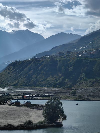 The image depicts a scenic view of a valley in Yamling, India, with a river in the foreground and mountains in the background. The river is wide and appears to be shallow, with a rocky bed visible in some areas. On the left bank of the river, there is a sandy area with a few small structures that appear to be huts or cabins. A larger body of water, possibly a pond or reservoir, is situated behind these structures. Further back, a row of tents or temporary shelters can be seen.
The middle ground of the image is dominated by a steep, green hillside dotted with trees and vegetation. A village is nestled on the slope of the hill, with several buildings visible. The buildings are mostly low-rise and appear to be made of stone or concrete.
The background features a series of majestic mountains, their peaks partially obscured by clouds. The mountains are layered, with the furthest ranges appearing hazy and blue due to atmospheric perspective. The sky is partly cloudy, with patches of blue sky visible between the clouds. The overall impression is one of a tranquil and picturesque landscape.