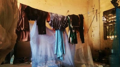The image depicts an indoor scene within a room in Rijiyar Zaki, Nigeria. An off-white or yellowish wall serves as the primary background, exhibiting areas of discoloration and wear. A clothesline is stretched horizontally across the upper portion of the room, from left to right. Hanging on this line are several items of clothing: a brown long-sleeved shirt, dark trousers, a patterned dark shirt with white stripes, a light blue fabric item, and a green and white striped garment. Clothespins are used to secure the laundry.

A large, light blue mosquito net is suspended from the clothesline in the central area, extending downwards and obscuring the lower background. A lighter, beige-colored mosquito net is partially visible on the far left. On the floor in the mid-left section, a light blue and white electric iron with a dark power cord is situated.

The right side of the room features a rectangular window secured with dark metal bars. Below the window, several metallic cooking pots and bowls are stacked on the floor or a low surface, adjacent to a tall, clear plastic container. A white utility box is mounted on the wall near the window. Between the window and the left half of the room, a light-colored, worn door or panel is visible on the wall. No individuals are present in the scene.