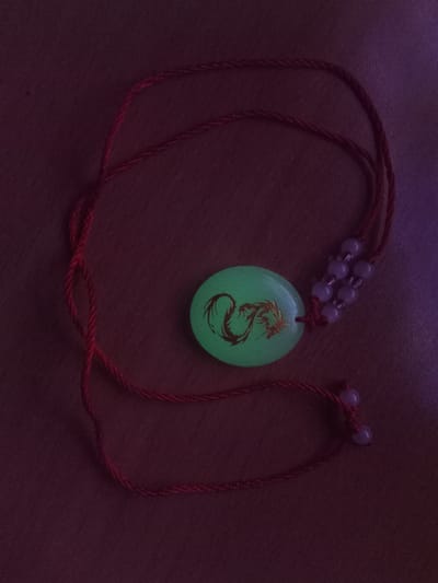 The image shows a close-up, overhead view of a necklace with a luminous green pendant featuring a gold dragon design. The pendant is oval and is strung on a thin red cord. Several small, light-purple beads are threaded onto the cord near the pendant, and two more beads are tied to the end of the cord. The necklace is lying on a dark, textured surface, possibly wood, which appears to be dimly lit and has a purple hue on the right side. The image's context does not provide any information about the city of Usman, Russia.