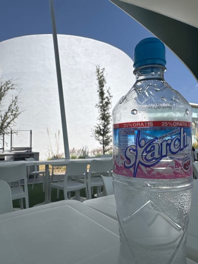 The scene depicts an outdoor setting in the city of Las Crucitas, Mexico, under a clear blue sky. In the foreground, a clear plastic bottle of "Spärch" natural water, containing 750ml, stands on a white, rectangular outdoor table. The bottle has a blue screw cap and condensation droplets are visible on its surface. The label is white, blue, and pink, prominently displaying "Spärch," "agua natural," and "25% GRATIS." A production code "L 100226 20:42 3 GDL CAD 10 FEB 27" is embossed on the bottle near the neck.
The midground shows additional white plastic tables and chairs arranged on an artificial green turf surface. To the left, a metal outdoor grill structure is visible. Several slender trees with sparse foliage and some tall, dry grasses are present in the background. A large, white, curved architectural canopy, supported by a grey vertical pole, dominates the upper background. No individuals are visible.