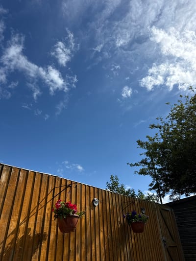 This outdoor image is an upward-angled view, predominantly featuring a bright blue sky filled with scattered white, wispy clouds across the top and left. A section of a dark green tree canopy extends from the top edge, its dense foliage partially obscuring the sky.

On the right side of the frame, a segment of a building's exterior wall is visible, constructed from horizontal wooden planks. The wood exhibits warm brown and orange tones, suggesting natural weathering. Attached to this wall are two terracotta-colored hanging flower pots. The upper pot displays vibrant purple flowers amidst green leaves, while the lower pot holds striking red flowers and foliage. Above each pot, a small, circular light fixture is mounted on the wooden surface. Strong, angled shadows, cast by the flower pots and tree branches, are visible on the wooden wall, indicating direct sunlight.

The scene captures a clear and sunny day, with the sun appearing to be high in the sky, suggesting it is either mid-morning or afternoon. There is no discernible text within the image. The setting appears to be a pleasant garden or outdoor space next to a wooden structure, consistent with a location such as Hazlemere, United Kingdom.