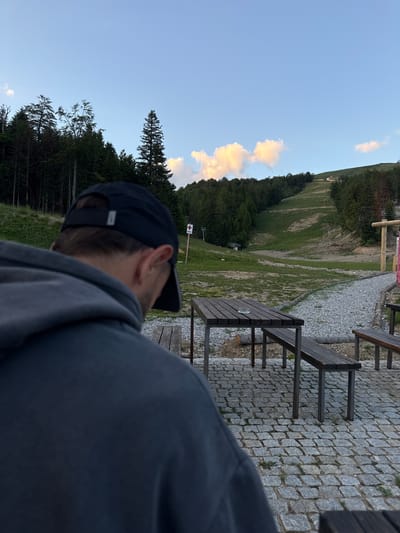 The image depicts an outdoor setting, possibly a rest area or a ski resort, with a person in the foreground wearing a dark gray hoodie and a black baseball cap, viewed from the back. They are facing a ski slope in the background, which is covered in green grass with a visible track. Trees line the sides of the slope, and the sky above is clear with some scattered clouds. In the mid-ground, there are wooden picnic tables and benches on a cobblestone patio. A red and white cross sign is visible on a pole to the left of the slope. The overall impression is of a serene, natural environment, likely in a mountainous region. The location is identified as Katun Bačko brdo, Montenegro.