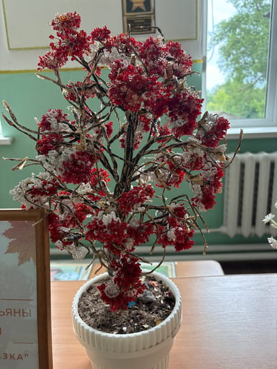 A close-up, eye-level shot captures a handcrafted tree adorned with red and white beads, resembling berries or flowers. The tree is planted in a white pot filled with soil and is situated on a wooden desk. To the left, a portion of a wooden frame holding a graphic with text is visible. In the background, a green wall with a framed certificate and a window revealing blurred green trees and a white radiator can be seen. This scene is likely located in an interior space such as a classroom or office within the Ветрынскі сельскі Савет region of Belarus. There are no people or visible actions depicted.