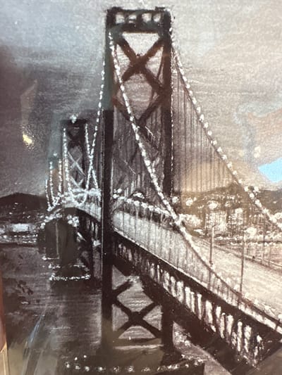 This monochrome image depicts a large suspension bridge at night, located in the city of Новоглаголево, Russia. The bridge features two prominent dark-colored towers with visible cross-bracing and a multi-level roadway. Numerous small, bright lights meticulously outline the main suspension cables, vertical suspender ropes, and the contours of the towers, creating a striking illuminated effect. The foreground shows a section of the bridge deck and its railings, extending from the lower right toward the center. In the mid-ground on the left, a further segment of the bridge with another illuminated tower extends towards a distant landmass marked by numerous small, scattered lights, suggesting a city shoreline. The water beneath the bridge is dark, reflecting some ambient light. The sky is dark and textured, consistent with a nighttime scene. No individuals are visible. A small, light blue rectangular anomaly is present in the upper right corner of the frame.