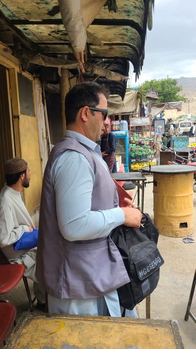 This image captures an outdoor scene in what appears to be a bustling market or roadside area in Kuchlak, Pakistan, under an overcast sky. The primary focus is on a man seated at a table or counter, viewed from the chest up and from his right side. He wears a light blue button-down shirt paired with a dark grey waistcoat and black sunglasses. His right hand holds a reddish-brown smartphone. To his left, a black backpack rests on the surface, clearly displaying the brand name "MOGESSIMO" in white letters.

Below and slightly in front of the main subject, another man is partially visible, wearing a light-colored shalwar kameez and a brown prayer cap (kufi), looking upwards. Red chairs are visible in the foreground, suggesting a seating area.

The background reveals a vibrant, informal market setting. Various stalls line the area, stocked with goods such as packaged snacks, drinks, and other sundry items. A large yellow barrel, possibly a tandoor oven or a storage unit, stands prominently to the right. Several motorcycles and other vehicles are parked further back. The market structures appear somewhat rustic, with makeshift canopies made of patterned fabric and tarpaulin providing shade. In the far distance, rugged mountains rise under the cloudy sky, hinting at the region's geography. The overall atmosphere suggests a casual daytime gathering or a brief stop at a local eatery.