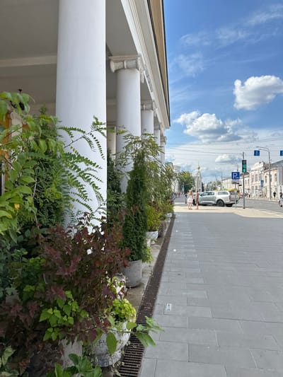 The image depicts a street scene in Golovinskoe, Russia, characterized by classical architecture and a sunny day. On the left, a colonnade of white columns lines the building, with potted plants, including green foliage and dark red leaves, placed in front of them. A drain grate is visible along the edge of the sidewalk.
The street itself is paved with grey rectangular tiles. A silver SUV is parked on the right side of the street, facing away from the camera. Two individuals, a man and a woman, are standing near the SUV, appearing to converse.
Further down the street, traffic lights are visible, along with road signs, including a blue "P" indicating parking. Buildings with light-colored facades line the street on the right. In the distance, a church with a golden dome stands prominently, suggesting a historical or religious site. Power lines and streetlights are also present. The sky is blue with scattered white clouds, indicating pleasant weather. The overall scene captures a moment of urban life with a blend of historical and modern elements.