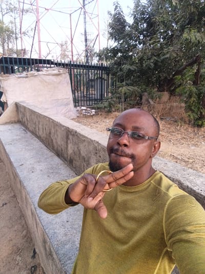 The image shows an adult male in an outdoor setting in Garun Gawa, Nigeria. The individual, with dark skin, a shaved head, and short facial hair, wears clear-framed glasses and an olive-green long-sleeved shirt. He is positioned in the lower right of the frame, facing the camera, with his right hand raised, displaying a gold-colored ring on his ring finger while forming a 'V' shape with his index and middle fingers. In the mid-ground, a light-grey concrete structure with two parallel tiers extends from the lower left. Behind this, a dark green metal fence is visible, with a light-colored tarpaulin or fabric-covered object directly behind it. The background features a large, reddish-brown metal lattice tower on the left against an overcast sky, and dense green foliage from trees and bushes on the right. The ground is dry and dusty with sparse vegetation.