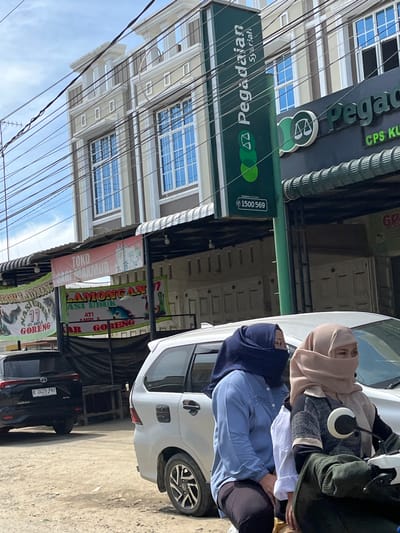 This is a vertical, eye-level, medium shot photograph taken in Kota Kuala Simpang, Indonesia. The scene depicts a street with several buildings and parked cars. In the foreground, the side of a white car is visible, and two women are seated inside it, wearing hijabs and traditional Islamic attire. The woman in the driver's seat appears to be wearing a blue hijab and blue long-sleeved shirt, while the passenger is wearing a light brown hijab and a patterned sweater. A black car is parked behind the white car.

Dominating the background are several buildings with multiple stories, featuring windows with blue panes. A prominent green sign with white text reads "Pegadaian Syariah," which is a Sharia-compliant pawnshop. Another sign above it, also in green, displays "Pegadaian" and "CPS KU." Below these, banners hang, one with pink text reading "TOKO SARI MAKMUR" and others with colorful graphics and text in Indonesian, such as "LAMONGAN" and "GORENG" (fried). Overhead, a complex network of wires crisscrosses the sky. The lighting suggests it is daytime, and the weather appears clear. The overall impression is of a busy urban street in Indonesia.