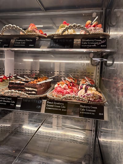 The image shows a display case containing several cakes and pastries, presumably in a bakery or cafe in Jianye District, China. The items are arranged on ornate silver trays with decorative beaded edges. From top to bottom, left side, the cakes are Strawberry Cream Cake Croissant and Strawberry Matcha Cream Cake, priced at ¥25 and ¥23. On the right side, there are Blueberry Cream Cake Croissant (¥25), French Strawberry Coconut Cream Cake (¥26), and Caramel Crunch Dark Chocolate Cake (¥17). The cakes are decorated with fresh fruits. Price tags are visible, showing the names of the pastries in English and Chinese, along with their prices in Yuan. The case is illuminated by vertical light bars.