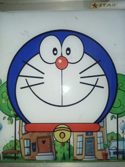 The image presents a detailed graphic of the cartoon character Doraemon, occupying the majority of the frame. Doraemon is depicted with a prominent blue outline around its head, a white face, two large white eyes with central black pupils, a small red circular nose, a broad black-lined smile, and three black whiskers extending from each cheek. Below the head, a red collar with a yellow bell is visible. The bell features concentric circles and a central dot.
The lower portion of the image, beneath Doraemon's head, reveals a stylized cartoon neighborhood. This background includes simplified depictions of houses, green trees and bushes, a brown park bench on the left, and a partially visible light blue vehicle on the right. One house on the left has a brown door with a heart-shaped window and the number "17" on its side, while another house has a blue door. The graphic is printed on a white, slightly reflective surface, which exhibits minor scratches and dust particles. A thin metallic frame borders the top edge. In the upper right corner, a "STAR" logo, featuring a red five-pointed star preceding the capitalized word "STAR," is present. No individuals, direct actions, or interactions are depicted. The specific location within the city of Inxuba Yethemba Local Municipality, South Africa, is not discernible from the visual content of the image.