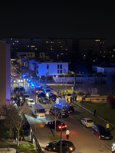 The image displays an elevated nighttime view of a street scene in Bron, France, centered around an apparent incident. The paved street is occupied by numerous vehicles, including several emergency vehicles with active flashing blue lights, specifically at least two police vans marked "POLICE" and a red fire truck. Approximately two dozen civilian vehicles, encompassing various sedans, hatchbacks, and vans, are parked or stationary along the roadway and adjacent sidewalks.
A significant gathering of people, estimated to be over fifty individuals, is distributed across the street, sidewalks, and a grassy park area situated to the right. Some individuals are identifiable as uniformed emergency personnel, while others appear to be civilians observing the scene. Streetlights provide primary illumination, casting shadows. Residential buildings with illuminated windows line the background, with one prominent, modern-style building displaying a strong blue luminescence, potentially from internal lighting or a reflection of emergency lights. The environment includes bare trees and patches of grass flanking the street. One individual in a white top and dark pants is observed walking towards the foreground, away from the central gathering, carrying a red object.
