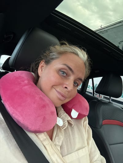 A woman with blonde hair and blue eyes, wearing a cream-colored shirt and a pink neck pillow, is sitting in a car with a sunroof. She is looking at the camera and smiling. The interior of the car is visible, including black leather seats with red accents and a black seatbelt. The image appears to be taken during the daytime. There is no indication of the specific location within the city of Ban Nong Bua, Thailand.