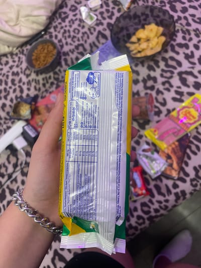 A person's hand with a silver bracelet is holding a food wrapper. The wrapper is green and yellow and has nutritional information printed on it in multiple languages. The background is a leopard-print fabric. There are several small packages of snacks scattered around the fabric. One of the snacks in the background appears to be a lollipop.
