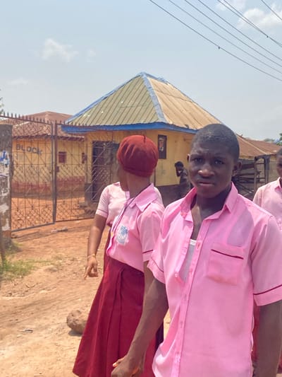 This image depicts an outdoor scene in Afikpo, Nigeria, under a bright, sunny sky with scattered clouds. In the immediate foreground, a young dark-skinned male, appearing to be a student, is positioned on the right, looking directly at the viewer with a neutral expression. He wears an unbuttoned light pink short-sleeved collared shirt over a white undershirt. To his left and slightly behind him, a young dark-skinned female, also appearing to be a student, is captured in motion, her back largely facing the camera but her head turned slightly to the left. She wears a dark red beret, a light pink short-sleeved collared shirt featuring a school emblem on the left chest, and a dark red long skirt. Her left arm is bent, revealing a thin light-colored bracelet. A third individual, partially visible on the far right, also wears a pink shirt.
The ground beneath the subjects is an unpaved, reddish-brown dirt path. In the midground, a prominent yellow single-story building with a distinctive pitched roof is visible. The roof appears to be constructed of corrugated metal, varying in color from light tan to olive, with a blue trim along its eaves. A dark metal gate with intricate scrollwork, behind which the word "BLOCK" is written in white on a wall, stands to the left of the main yellow building. Another blurred, dark-skinned figure is observed leaning or bending near the central yellow building to the right. Utility lines stretch horizontally across the upper portion of the frame against the sky.