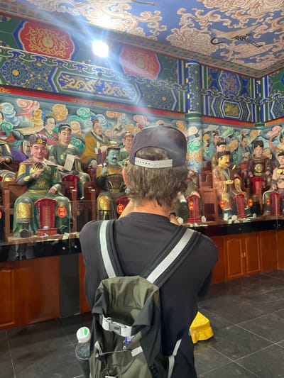 A person wearing a grey baseball cap and a black sweatshirt stands inside a building, possibly a temple or cultural site, in Lintong District, China. The walls are decorated with colorful murals, including images of figures in traditional Chinese clothing. The walls could be decorated with statues. The floor is dark and tiled. The person is wearing a backpack.