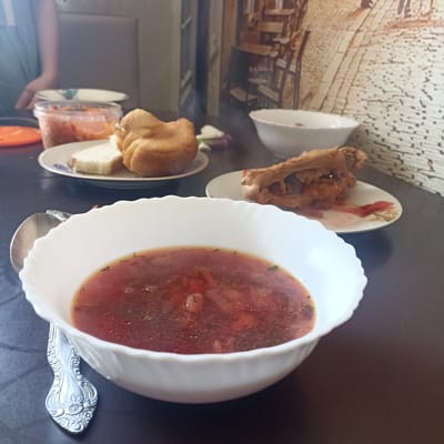 A table is set for a meal, with a bowl of borscht in the foreground, to the right of a decorative spoon. The borscht is a rich red color with visible pieces of vegetables and herbs. To the left of the borscht, a plate holds a slice of white bread and a golden-brown, round bread roll, likely a pampushka. Further back on the table, another plate contains a piece of fried chicken. A clear plastic container filled with orange shredded vegetables, possibly carrots or kimchi, is also visible. The table is dark brown, and the background shows a wall with a patterned wallpaper depicting a scene that appears to be a street with buildings and chairs. A portion of a person's arm and shoulder, wearing a blue garment, is visible on the left side of the image, suggesting a person is seated at the table. The lighting appears to be indoor, possibly natural light from a window, suggesting it could be daytime. The overall impression is of a home-cooked meal being prepared or enjoyed. No visible text is present in the image. The scene is set in Shakhunya, Russia.