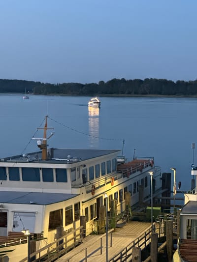 The image depicts a twilight scene at the harbor of Prien am Chiemsee, Germany. A large passenger boat is docked at a wooden pier in the foreground. The boat is illuminated by interior lights and external spotlights, casting a warm glow on the water and the wooden planks of the pier. The boat has multiple decks and numerous windows. A smaller boat is visible further out on the lake, and another larger boat with lights is moving away from the viewer. The background is dominated by a dark, tree-lined shore. The sky is a clear, deep blue, indicative of dusk. The scene is tranquil and serene, with the gentle lapping of water and the distant hum of engines being the only suggested sounds.