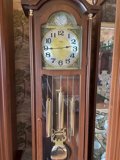 A close-up, horizontal shot shows a grandfather clock made of dark wood. The clock face is a silver circle with black Arabic numerals and gold accents. The clock has a second hand and a minute hand, which is pointing to the 12. The hour hand is pointing to the 3. To the right of the clock face, a glass-enclosed section reveals three brass pendulums hanging from chains. The pendulums are polished and reflect the light. Above the clock face, a shelf displays several crystal glasses and cups. The background is a wall with a repeating pattern of pine trees. The scene appears to be indoors, with soft lighting. There is no indication of the time of day or weather. No people are visible. The location context provided is Krasnoe-na-Volge, Russia, but there are no visual cues in the image to confirm this. Visible text on the clock face includes "SUIK" and numbers 1 through 12.