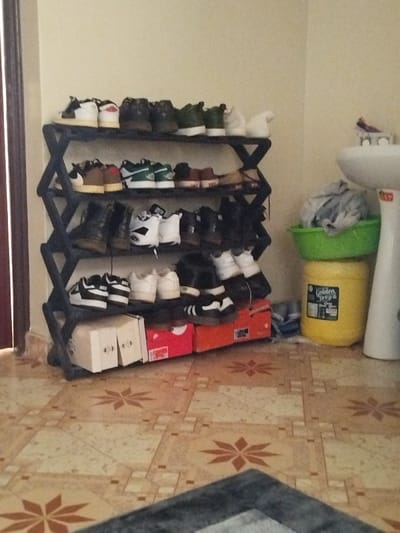 The image shows a black shoe rack with four shelves, filled with a variety of shoes. The rack is located in a room with tiled flooring, and there is a white porcelain sink and a yellow container labeled "Golden Drop" to the right of the rack. The room is likely an entryway or a hallway, and the shoe rack is placed against a light-colored wall. The floor has a beige and brown tiled pattern.