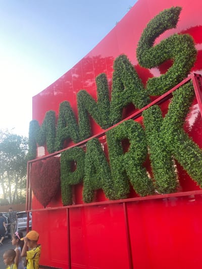 The image depicts a vibrant red wall adorned with large, three-dimensional letters formed from lush green bushes, spelling out "MANAS PARK". To the left of the letters, a large, red, heart-shaped shrub is visible. The letters are positioned on a red frame structure. In the foreground, three young children are present. The child on the left is a baby with a shaved head. The middle child is a girl with a pink bow in her hair, wearing a white shirt. The child on the right is a boy wearing a yellow and blue soccer jersey and a yellow cap, looking upwards. Behind the children, a man in a patterned shirt and beige pants is walking away from the camera, and a silver car is parked. The background features trees and a clear sky. The overall scene suggests an outdoor setting, likely in a park or recreational area.