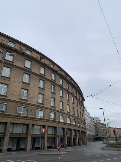 The image shows a curved, stone-faced building with numerous windows, located on a street in Frankfurt am Main, Germany. The building is likely a residential building and the street is empty of pedestrians and vehicles. Several power lines run across the sky, which is grey and overcast. The building is a prominent feature of the scene, and its curved shape suggests a design from the mid-20th century. The building is surrounded by other structures, including a building on the right side of the image which is partially obscured by a row of trees. A street sign and a large white traffic sign with a red border are present in the foreground of the image. This street corner is located at the intersection of two roads, one with a tram line in the middle of the street. The buildings are in a state of good repair, with no visible signs of damage or neglect. The photo was likely taken during the day, as the lighting conditions suggest an overcast sky, and the buildings appear to be in a state of normal use. The image captures a typical street scene in the city of Frankfurt am Main, Germany, showing the mix of old and new architecture, the presence of power lines, and the overall feeling of a functional urban environment.