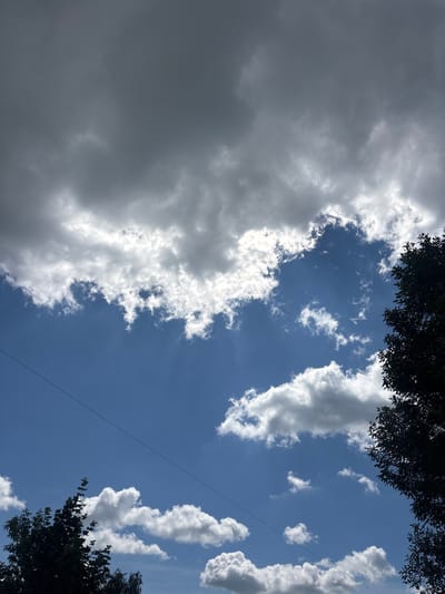 This media file depicts a cloudy sky with patches of blue visible. The clouds are varied, with some appearing dark and heavy, suggesting impending rain or overcast conditions, while others are bright white and fluffy, catching the sunlight. The sky is framed by the dark silhouettes of tree branches at the top and bottom edges, indicating an outdoor setting. A thin, dark line, possibly a power line or a thin branch, bisects the sky diagonally. The scene evokes a sense of transition, possibly between fair weather and a storm. The presence of trees and the quality of light suggest it might be daytime, perhaps late morning or afternoon. No people or specific objects are identifiable beyond the natural elements. The location context suggests this is a view of the sky over Pereiaslavske, Ukraine. The overall mood is somewhat dramatic due to the contrast between the dark and light clouds.