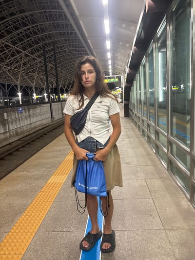 A young woman stands on a train platform in Gyeongju-si, South Korea. She is wearing a white collared shirt, a denim skirt, and a tan skirt. A dark blue fanny pack is slung across her chest. She holds a blue drawstring bag with "Google Gemini" printed on it in her hands. The platform has a yellow tactile paving warning strip. In the background, train tracks are visible, as well as a large, covered roof structure and glass-paneled walls. The scene appears to be at night, with artificial lighting illuminating the platform.