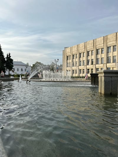 The image captures an outdoor scene featuring a large public fountain with water spraying upwards, set against a backdrop of a light-colored, multi-story building with numerous windows. The fountain is positioned in the foreground and midground, with rippling water surface. Several people are visible in the background, some near the fountain and others walking or sitting. To the left of the fountain, a white building with a cross on its roof is partially visible, along with some trees. The sky is partly cloudy. The overall impression is of a public square or park area in a city.