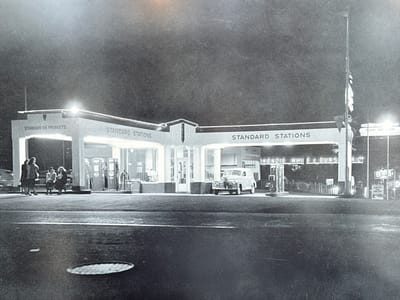 The photograph captures a Standard Oil station in Langley, Canada, likely around the mid-20th century based on the vehicle and clothing styles. The station features a multi-section canopy with "STANDARD OIL PRODUCTS" and "STANDARD STATIONS" prominently displayed. Several gas pumps are visible under the canopy. A vintage panel truck is parked near the right side of the station. Behind the station building, additional signage reads "KENZIE WHITE & DUNS RLTO". The surrounding area includes signs for other businesses, including one for "BA" (British American Oil). On the left side of the station, a group of four women are sitting on a bench. The ground is paved and has a manhole cover visible. The scene is illuminated by lights from the station and nearby signs, creating a contrast with the dark sky.