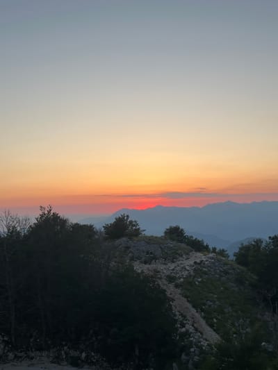 This image depicts a mountain landscape at sunset, likely from a high vantage point in or near Kuk, Montenegro. The sky transitions from a pale blue at the top to a vibrant orange and red band near the horizon, where the sun is setting behind a range of layered mountains. In the foreground, a rocky, tree-covered slope is visible, with a worn path winding its way up. The trees are dark green, with some bare branches suggesting a cooler season or a natural mountainous terrain. The overall atmosphere is serene and vast, capturing the beauty of the natural environment at dusk.