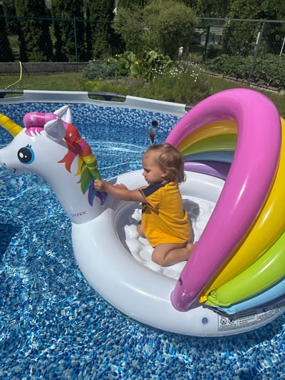 A young child with blond hair pulled into two small ponytails, wearing a yellow t-shirt with navy accents and no pants, kneels inside a white, inflatable unicorn pool float in a swimming pool. The unicorn float has a rainbow-colored mane and horn. The pool water is blue with a patterned liner. The background shows a green lawn, a dark fence, and lush green foliage, suggesting an outdoor residential setting. The image is taken from a slightly elevated, side angle, focusing on the child and the float. The location is identified as Astrashitskagaradokski rural council, Belarus.