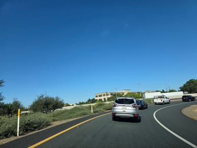 The image depicts a bright, sunny day with a clear blue sky, suggesting it is mid-morning or afternoon. The scene is a highway in Elk Grove, California, with cars traveling along a paved road. The road has a yellow lane marker and a white dividing line. On the left side of the road, there are green bushes and trees, along with a yellow and white reflective marker post. In the distance, there are several buildings, appearing to be offices or a business park.
Several vehicles are visible on the road. Closest to the viewer is a silver SUV, followed by a dark-colored sedan. Further ahead, there is a white pickup truck with a rack on its bed, and another dark SUV. The vehicles appear to be in moderate traffic, moving along the highway.
One of the visible license plates on the silver SUV reads "8TGN694". The overall impression is a typical drive on a sunny day in a suburban or commercial area. There are no apparent signs of urgency or distress; the scene is calm and ordinary. The clear sky and bright sunlight indicate good weather conditions.