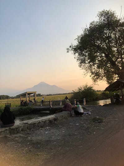 This is a medium shot of a rural landscape in Keboguyang, Indonesia, at dusk. The sky is a soft gradient of pale blue to pale orange. A large, hazy mountain dominates the background, with its peak just visible. In the middle ground, a small, open-sided shelter with a corrugated metal roof is supported by wooden posts. Several motorcycles are parked under the shelter, and two people appear to be inside. A narrow bridge spans a small canal or stream in front of the shelter. On the bridge, a person stands holding a stick, and further across, several other figures are visible. In the foreground, a concrete retaining wall runs along the edge of the canal. A person wearing a black headscarf and a purple skirt sits on the wall facing away from the camera. To their right, a person wearing a white t-shirt and a green cap squats on the ground, while another person in a patterned dress sits beside them. A blanket is spread out on the ground near the squatting figure. The ground is unpaved, with dirt and sparse vegetation. A large tree with dense foliage is in the upper right corner of the frame, partially obscuring the sky. To the right of the canal, there is a small, rustic structure that appears to be another shelter. The overall scene depicts a peaceful, everyday moment in a rural Indonesian setting as the day ends.