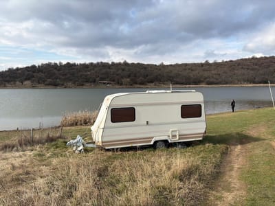 A white and beige caravan is parked on a grassy bank adjacent to a calm body of water, likely a lake or reservoir, under an overcast sky near Cherna Gora, Bulgaria. The foreground features dry, yellowish-brown grass, with a dirt path visible on the right leading towards the water. A single individual, clad in dark clothing, stands near the water's edge on the right side of the frame, facing away from the camera. Fishing equipment, including a rod on a stand, is positioned on the grass near this person. Items covered with a light-colored tarp are situated on the ground near the caravan's hitch. A wire fence runs along the left side of the bank. In the background, a treeline of bare deciduous trees covers a gentle slope across the water, with a small structure faintly visible within the trees on the far bank.