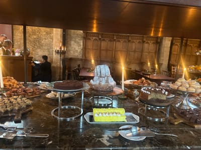 The image displays an indoor dessert buffet within a large, historic room located in Cernay-la-Ville, France. The room's background features stone block walls and sections of dark wooden paneling with carved details. The lighting is predominantly warm and dim, emanating from multiple lit taper candles in silver holders on the buffet and candelabras in the background, creating a historic atmosphere.
In the foreground, a dark, polished marble-effect countertop serves as the buffet table. It is laden with various desserts: a round dark chocolate cake on a silver stand, a tall conical fluted cake dusted with powdered sugar on a wooden board, a light-colored cake with white frosting and floral decorations (partially sliced) on another silver stand, and a tiered silver stand holding multiple sugar-dusted round pastries (donuts). Other items include a rectangular yellow cake with meringue swirls on a white platter, numerous small pastries, muffins, and tarts arranged on wooden boards and platters. Silver serving tongs and spatulas are provided on small white plates.
In the left background, two individuals are partially visible near a dark wooden counter; one male figure in a vest and shirt interacts with another. Further into the mid-ground, a person dressed in dark attire stands with their back to the viewer, operating a machine or device on a stand. The deeper background reveals additional dark wooden tables and chairs, with one table draped in a dark cloth, suggesting an event or dining area.