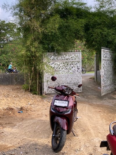 In Cottrueng, Indonesia, a maroon Honda electric scooter is parked on a dirt path in front of an open, ornate white metal gate. The gate is flanked by dense green bamboo foliage, which forms an archway over the entrance. In the background, beyond the gate, pink flowers are visible, and a person on a motorcycle is seen on a paved road. The ground is covered with loose dirt, stones, and some scattered debris. Another red scooter is partially visible in the lower right corner of the image. The sky is overcast.
