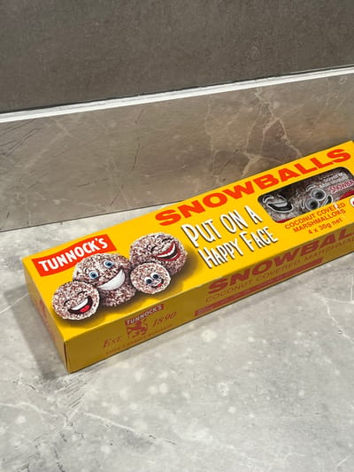 A yellow box of Tunnock's Snowballs is displayed diagonally on a grey and white tiled surface. The box features cartoon snowballs with smiling faces. Text on the box reads "SNOWBALLS," "PUT ON A HAPPY FACE," "COCONUT COVERED MARSHMALLOWS," and "TUNNOCK'S EST. 1890." The box indicates it contains 4 x 30g of product. A small transparent window shows the actual snowballs inside the box.