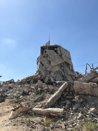 This image depicts a large pile of rubble in the city of Jabalia, Palestinian Territory, under a clear blue sky. The central feature is a substantial, irregularly shaped mound of fragmented concrete, twisted rebar, and other building debris, rising prominently against the horizon. This structure, likely the remains of a multi-story building, shows fractured concrete slabs leaning against each other, exposing internal reinforcement bars. The ground surrounding the main debris pile is also covered in smaller pieces of rubble, dirt, and sparse patches of green and yellow vegetation. No individuals or specific activities are visible in the scene.