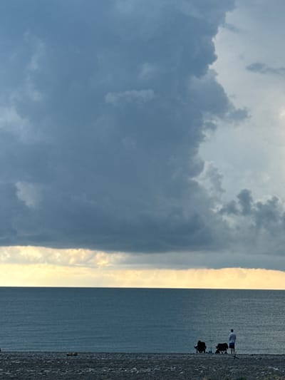 This image depicts a serene beach scene on the coast of the Black Sea, likely in Abkhazia, under a dramatic and cloudy sky. The foreground is a pebble beach, with two dark chairs set up, suggesting a waiting or resting posture. A single individual stands facing the water, dressed in a white shirt and blue shorts, with another person seated in one of the chairs. The sea is calm and stretches out to the horizon, meeting a strip of soft yellow and white clouds near the waterline. Above this, the sky is dominated by large, dark, and textured storm clouds, creating a striking contrast with the brighter sky on the right side of the frame. The overall mood is tranquil, with a sense of anticipation or contemplation due to the weather.