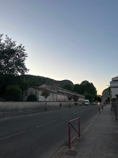 The image depicts a street scene in Anduze, France, likely during twilight. On the left, a long, low-rise building with a grey, weathered facade runs parallel to the road. It appears to be an industrial or commercial structure, with a fence running along its front. Trees and dense foliage are visible behind the building and on a rocky, tree-covered hillside in the background. The road in the foreground is asphalt, with white lane markings. A paved sidewalk runs along the right side of the road, constructed with cobblestones. Red metal railings are placed intermittently along the edge of the sidewalk. Several vehicles are present on the road, including a white van and two dark cars. The sky is a clear, pale blue, transitioning to a lighter hue towards the horizon, suggesting the setting or rising sun. The overall impression is one of a quiet, somewhat dated urban environment bathed in soft, natural light.