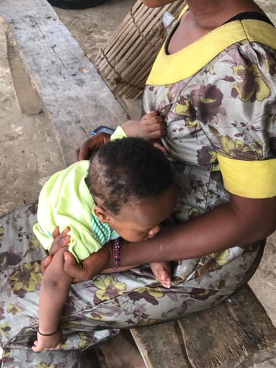 Overhead shot of a woman holding a baby in her lap. The woman wears a dress with a floral pattern and yellow trim, along with a beaded bracelet. The baby is dressed in a light green shirt. They sit on a weathered wooden surface, possibly a bench, with a cylindrical wicker item nearby. The ground is a light, earthy tone. The focus is on the intimate moment between the woman and child.