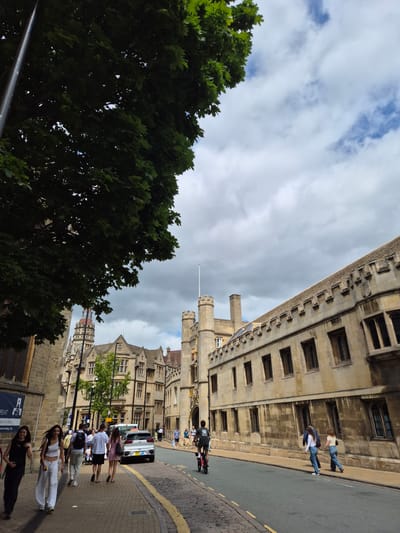 The image depicts a street scene in Cambridge, United Kingdom, likely near the University. On the right side of the street, a long, tan stone building with numerous rectangular windows and a crenellated roofline dominates the view. To the left, another stone building, appearing older and more ornate with a distinctive tower and clock face, is visible. A large, leafy green tree partially obscures the sky and upper portions of the buildings on the left.
The street itself is paved with cobblestones near the sidewalk and asphalt in the road. A white car is parked on the street. Several individuals are walking on the sidewalk. In the foreground, two women are walking towards the viewer, one in a black sleeveless top and dark pants, the other in a white tank top and white pants. Behind them, a group of people are walking away from the camera, including a man in a white t-shirt and black shorts, a woman in a pink dress, and another person in a white t-shirt and dark pants. Further down the street, more pedestrians are visible, as is a person riding a pink electric scooter. The sky is partly cloudy, with a mix of white clouds and patches of blue. A flag pole is visible on top of the building on the right. A blue sign with white text is posted on the wall of the building on the left.