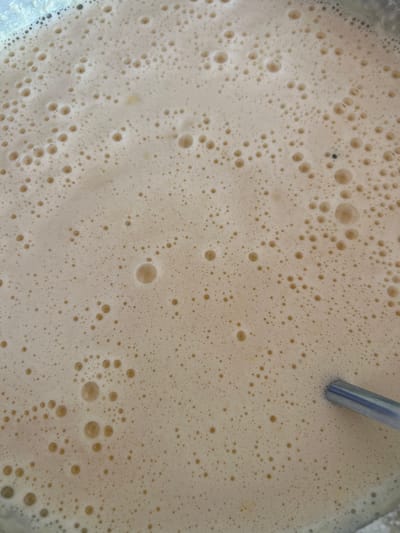 The image shows a close-up, top-down view of a light brown, foamy liquid with many small bubbles. A silver spoon is partially visible in the lower right corner, submerged in the liquid. The liquid appears to be in a container, with the edge of the container visible in the upper left corner. There is no indication of the location in the city of Üsharal, Kazakhstan within the image itself.