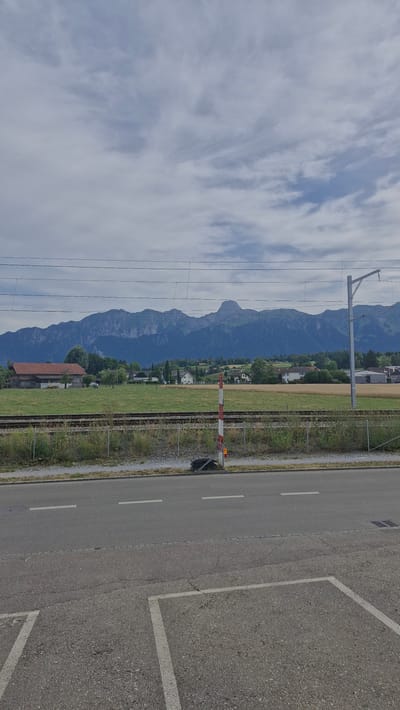 This image depicts a rural landscape in Uetendorf, Switzerland, under a cloudy sky, suggesting it is daytime. The scene is characterized by a juxtaposition of natural and man-made elements. In the foreground, a paved road with white lane markings runs diagonally across the frame, with a parking lot demarcated by white lines visible on the right. Adjacent to the road is a railway track with overhead power lines, indicating an active train route. A red and white striped railway crossing barrier is visible, suggesting trains may be passing or about to pass.
Behind the railway line, a green field stretches out, dotted with trees and a few houses, leading towards a backdrop of majestic, dark blue mountains. The mountains are imposing and appear rugged, partially obscured by the overcast sky. The presence of houses and cultivated fields suggests a settled, agricultural, and residential area.
The overall mood is tranquil and somewhat serene, with the dramatic mountain range providing a sense of scale and natural grandeur against the more mundane elements of the road and railway. The time of day is difficult to pinpoint precisely due to the cloud cover, but the lighting suggests it is not early morning or late evening. No people are visible in the image, and no text is discernible.