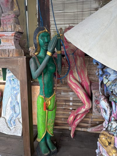 This image depicts an outdoor setting with several colorful, carved wooden statues. In the foreground, a green female figure stands with hands clasped in front of her chest and wearing a green sarong and a beaded necklace. To her right, a pink and gold abstract human form is visible, partially obscured by a white umbrella. To the left of the green figure, a carved white relief of a figure is leaning against a wooden structure. The background features a bamboo blind and other carved wooden objects, suggesting an artistic display or workshop. The scene is located in Tejakula, Indonesia, likely an outdoor area associated with local crafts or tourism.