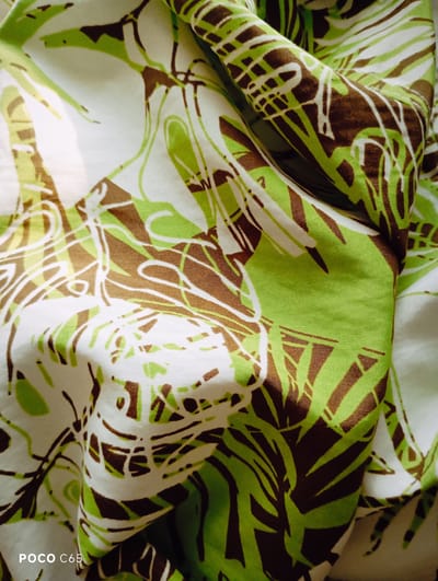 The image is a close-up, high-angle shot of a piece of fabric with a green, brown, and white abstract pattern. The fabric is draped and creased, creating shadows and highlights that add depth to the image. The pattern consists of organic shapes and lines, resembling stylized leaves or brushstrokes. There are no people or specific objects identifiable in the scene. The image is cropped to focus solely on the texture and pattern of the fabric. The location in the city of Zhytomyr, Ukraine, is not discernible from the visual content of the image.