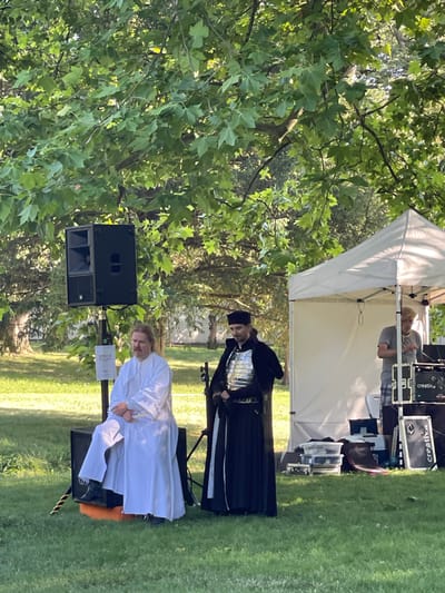 Two men are standing in a grassy outdoor area with trees and a white tent. One man, on the left, is wearing a long white robe and sitting on a black speaker. He has light-colored hair and a mustache. The second man, on the right, is wearing a black robe with a silver chest plate and a black hat. He is standing and holding a black staff. Behind him, under the tent, a third person in a gray t-shirt and plaid shorts appears to be operating some audio equipment. Several audio speakers, cases, and plastic storage containers are visible on the ground. A sign with the logo "EOE" is attached to a pole near the speaker. The scene appears to be set up for an outdoor event, possibly a performance or presentation, in the city of Stupava, Slovakia.